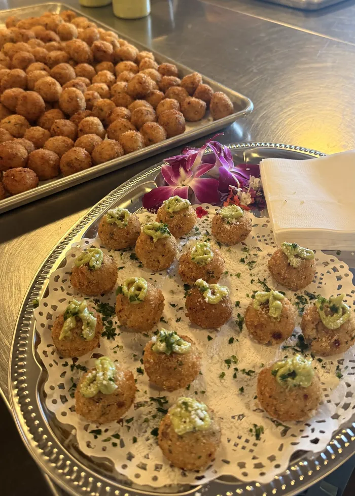 A silver tray of passed arancini topped with green sauce and parmigiano, with more trays of arancini behind